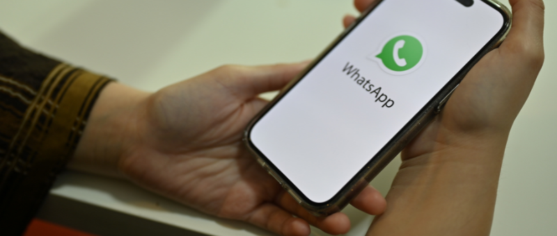 A imagem mostra uma pessoa segurando um smartphone com o aplicativo WhatsApp na tela. A pessoa está usando uma camisa preta com listras, e está sentada em uma mesa. O fundo da imagem é branco e o smartphone está em um ângulo para que o aplicativo seja visível.