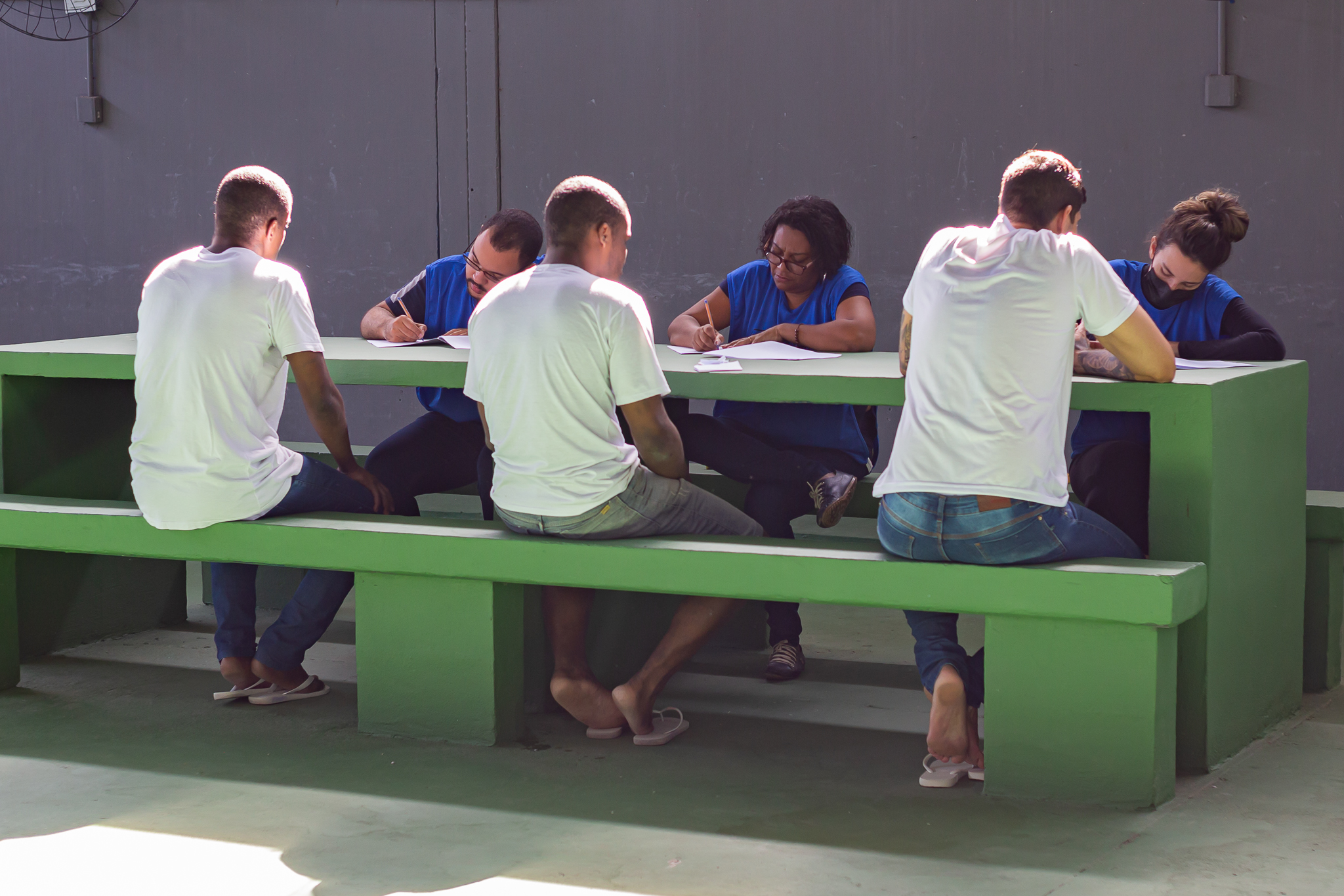 Detentos de camisas brancas sentados em bancos verdes recebendo atendimento de integrantes da equipe do programa Justiça Itinerante