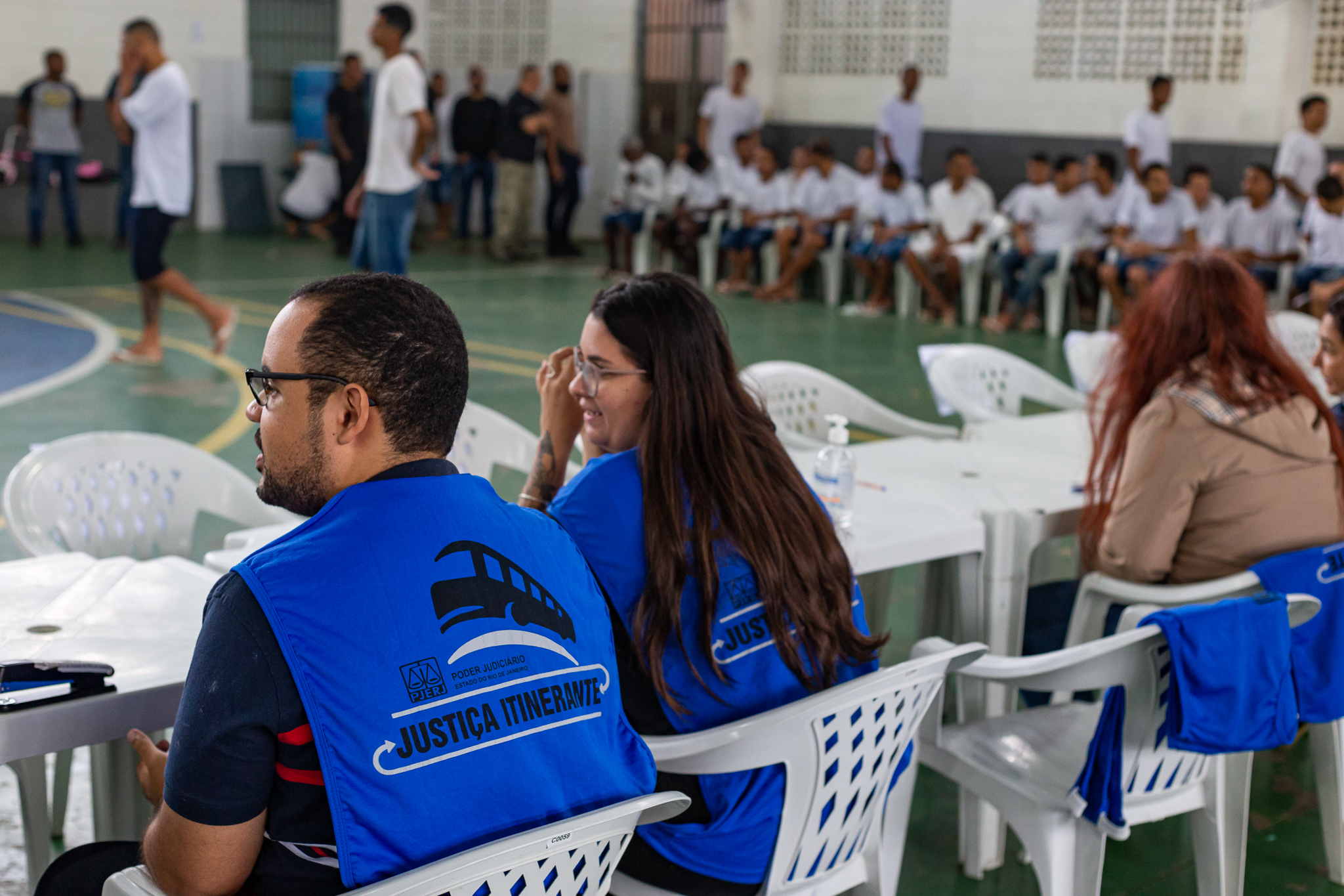 De costas, três integrantes com coletes azuis do projeto Justiça Itinerante sentados. Ao fundo, presos desfocados sentados aguardando atendimento 