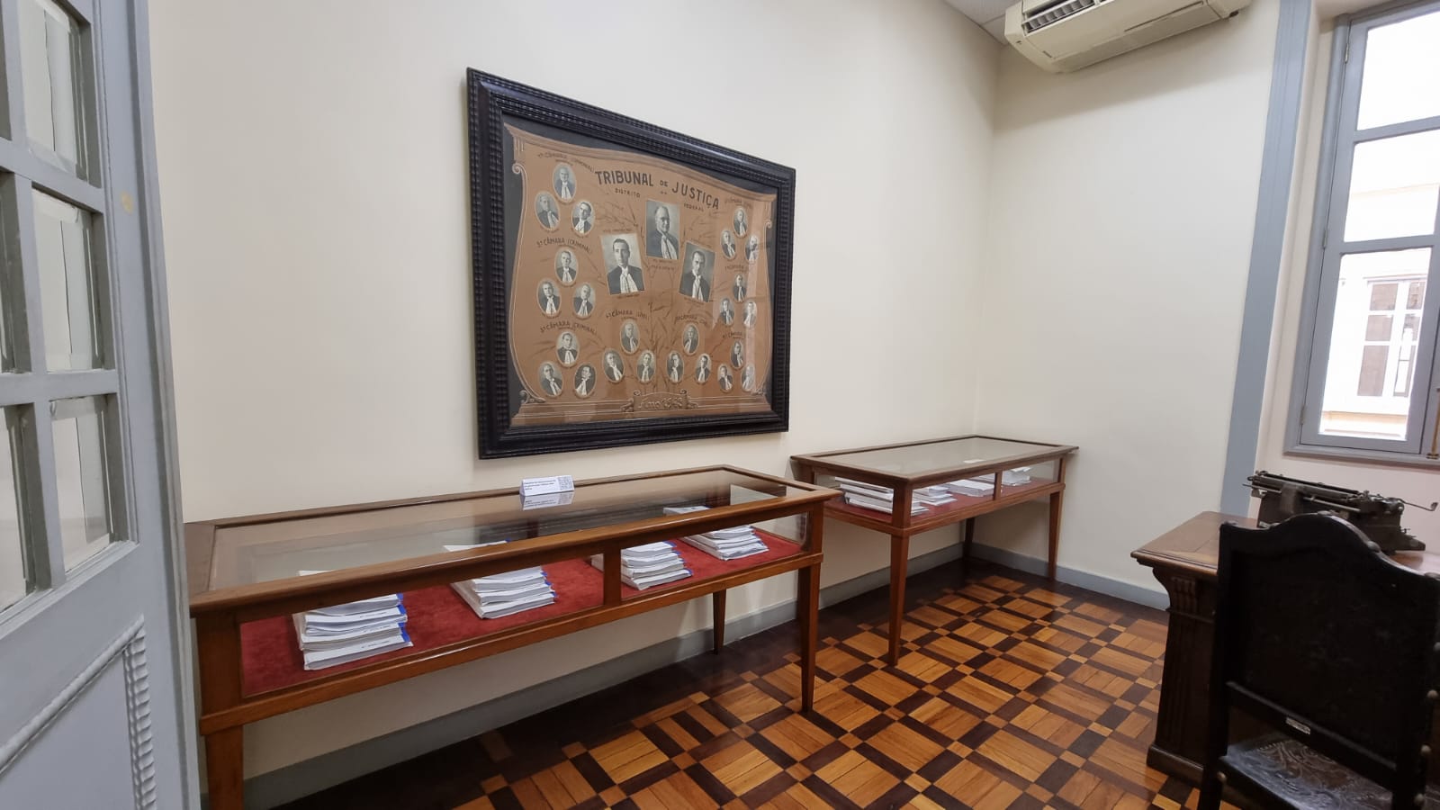 uma foto da sala do museu da justiça com duas vitrines horizontais revestidas de vidro. No interior delas, peças processuais. Na parede, um quadro ilustrativo com fotos de magistrados