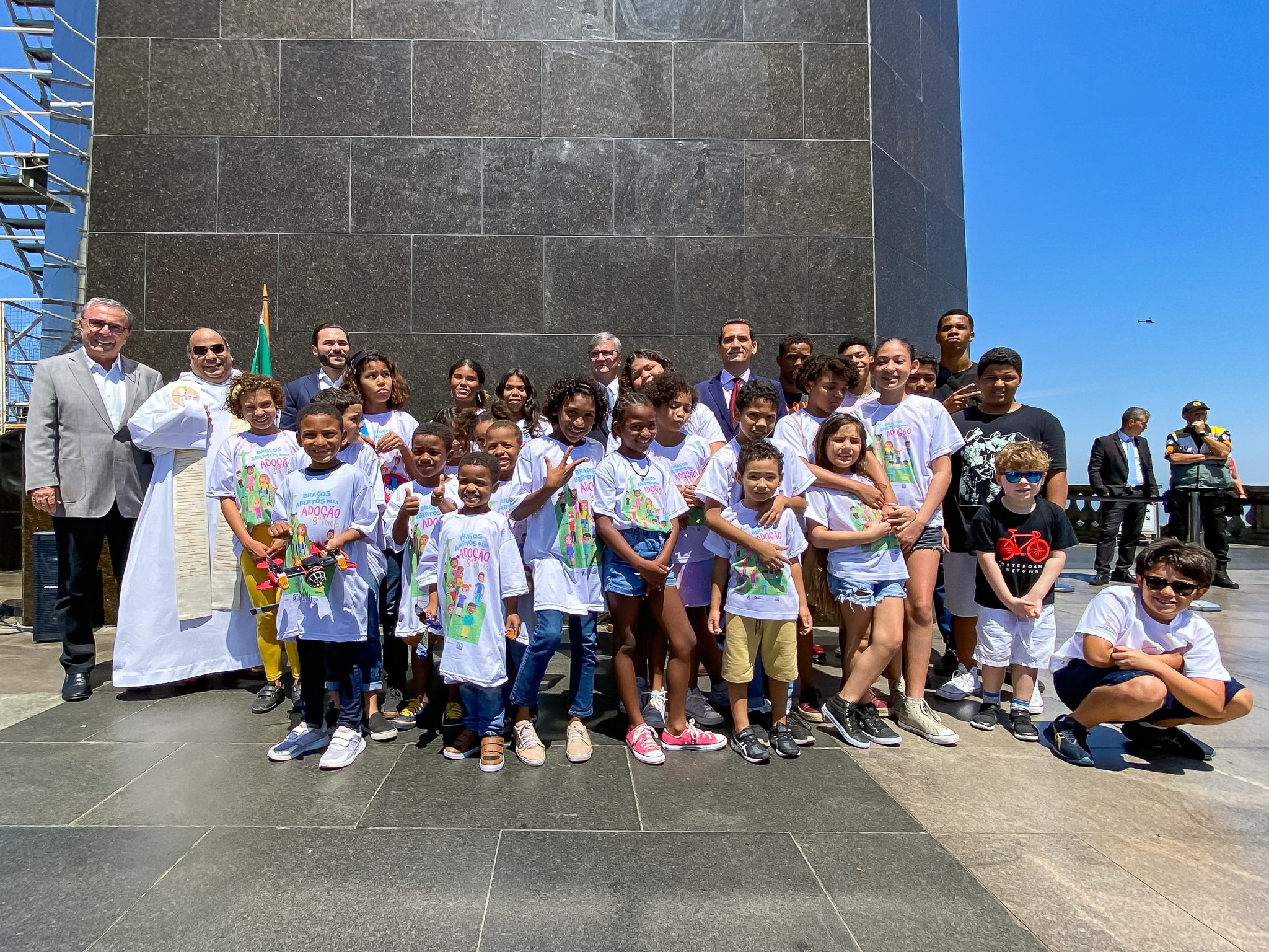 Foto posada: grupo de crianças e adolescente, acompanhados de autoridades aos pés do Cristo Redentor. Ao fundo céu azul. 