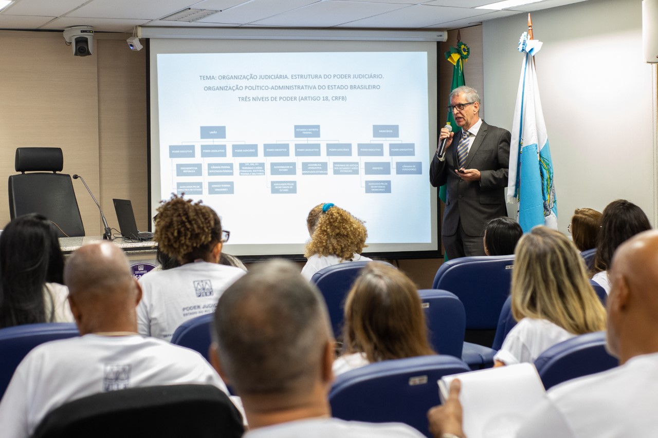 Na foto, o desembargador Heleno fala aos participantes do curso. Ele tem um microfone nas mãos e está de pé. A foto mostra as costas dos participantes, que estão sentados.