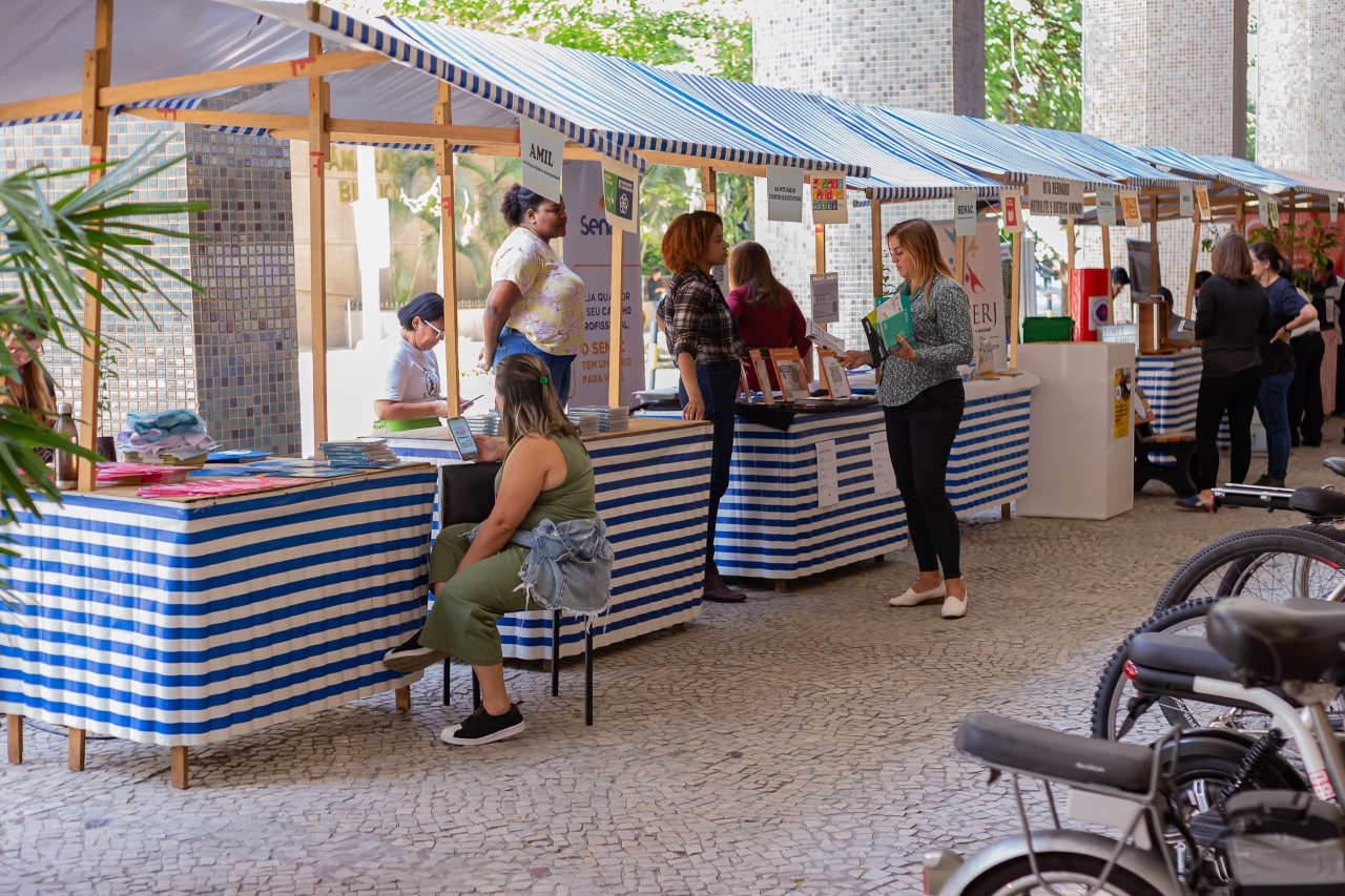 A imagem mostra as barraquinhas da Feira de Sustentabilidade, montada próximo à saída do térreo do TJRJ. Na fotos, visitantes recebem orientações dos expositores sobre meio ambiente e sustentabilidade