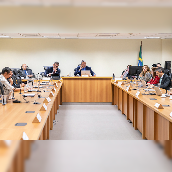 A imagem mostra uma reunião formal . A disposição é em formato de “U”, com mesas de madeira clara alinhadas dos dois lados e uma mesa central ao fundo, onde se encontra o desembargador Marcelo Anátocles que conduz a reunião. Todas as pessoas estão sentadas, a maioria com laptops ou documentos à frente.  À direita da imagem, uma bandeira do Brasil está posicionada no canto, 