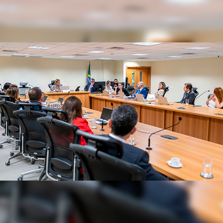 A imagem mostra um grupo de pessoas sentadas ao redor de uma mesa de madeira em uma sala de reunião 