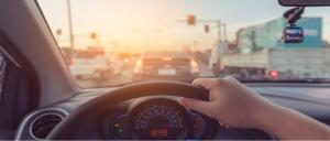 Imagem do trânsito intenso de uma rodovia ao fundo e, em primeiro plano,  visualizamos a mão do condutor ao volante e parte do painel do carro.