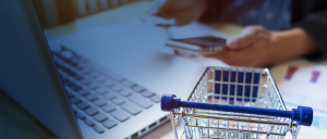 #ParaTodosVerem: Pessoa branca, de traje em tons de azul, sentada a uma mesa, sobre a qual estão um laptop e uma miniatura de carrinho de compras de supermercado, portando, na mão direita um cartão, e, na esquerda, um celular. A foto simboliza o consumo de produtos pela internet.