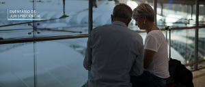 Imagem de um casal de costas, com uma mala, no saguão de um aeroporto, ao fundo algumas aeronaves estacionadas.