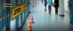 #ParaTodosVerem: Acima, no canto esquerdo, o título “DECISÃO”. À direita, pessoas indistintas no corredor de um local fechado. À esquerda, cones em frente a uma porta de vidro com lacre amarelo e preto contendo os dizeres “CUIDADO, EM OBRAS”.