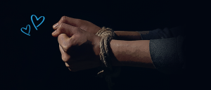 Imagem de fundo escuro com os punhos de uma pessoa amarrados com uma corda. Ao centro, na parte superior, um coração com a frase: em apoio a campanha coração azul do UNODC. 