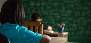 Imagem de um aluno sentado em uma carteira de sala de aula, com um lápis na mão 