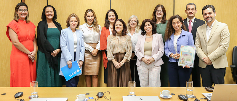 Foto mostra 12 pessoas, sendo dez mulheres, vestidas com diferentes roupas, e dois homens, um trajando terno escuro e outro de paletó bege e sem gravata, todos juntos, de pé, posando para a foto, sorrindo, tendo à frente uma mesa de madeira clara com notebooks sobre ela. Ao fundo, uma parede recoberta com chapas laminadas de material no mesmo tom da mesa