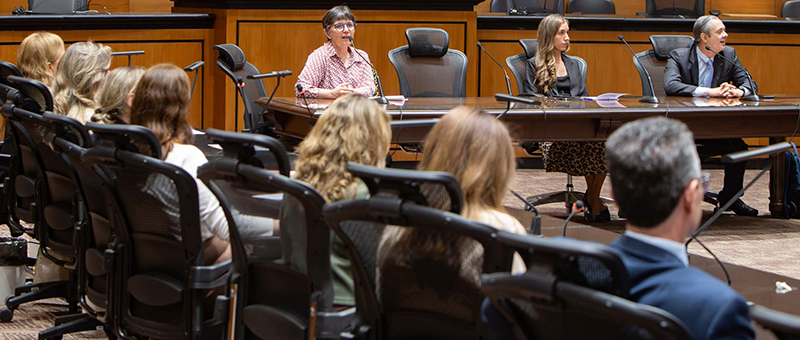 A imagem mostra uma palestra ou reunião realizada em um auditório institucional com acabamento em madeira. Em primeiro plano, participantes estão sentados em cadeiras voltadas para uma mesa de debates posicionada ao centro do espaço. Na mesa principal, três pessoas participam da apresentação. Uma mulher, ao centro, fala ao microfone enquanto os demais acompanham a exposição. O ambiente conta com cadeiras ergonômicas, púlpitos e microfones distribuídos pelo plenário.