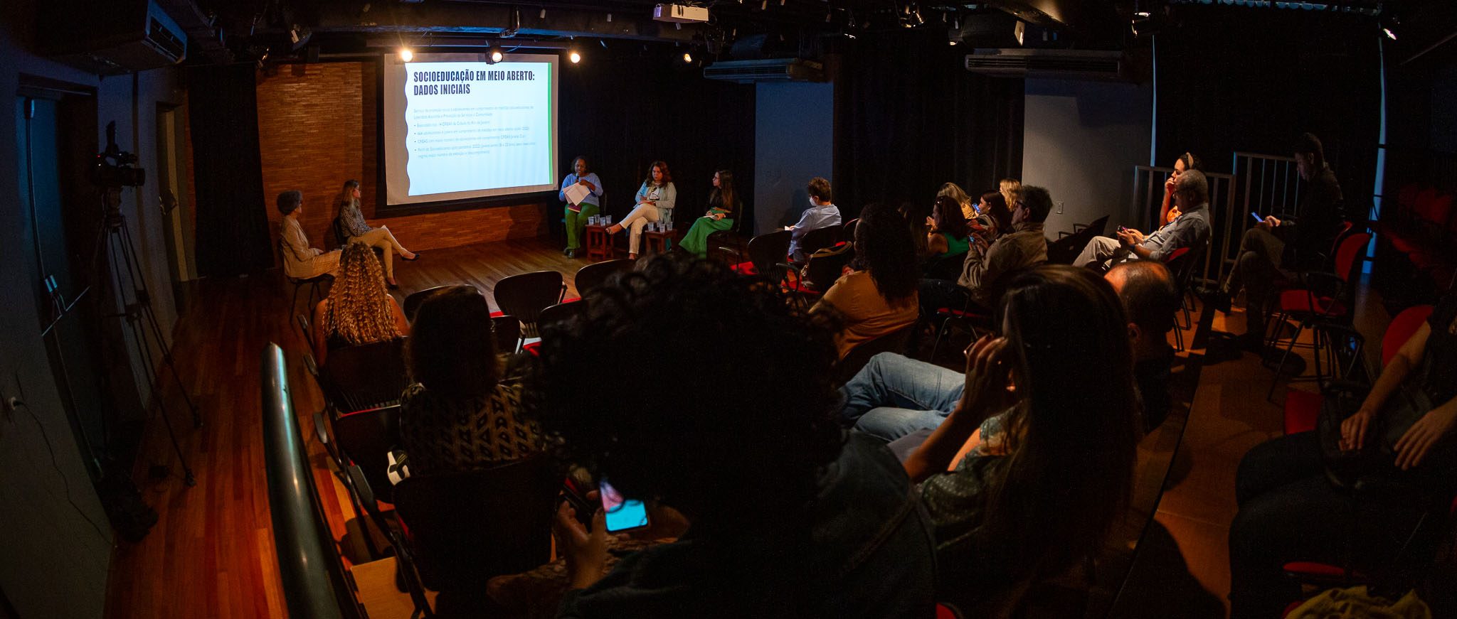 Imagem feita de uma sala mostrando os participantes do evento assistindo ao evento, com palestrantes e projeção de imagem ao fundo