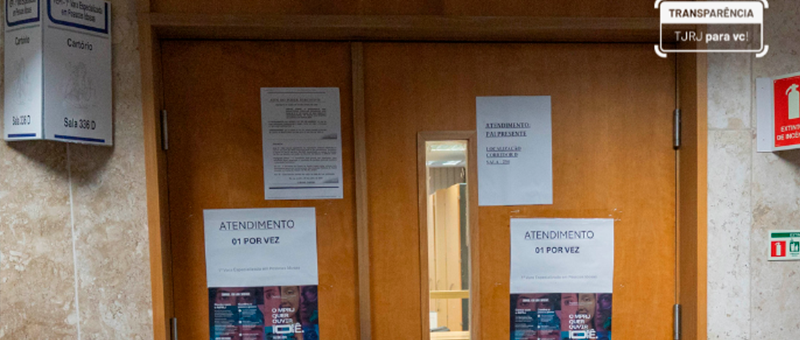 A imagem mostra uma porta dupla de madeira em um prédio público, com vários avisos impressos colados. Os cartazes informam regras de atendimento, como “atendimento 01 por vez”, e há sinalização institucional do tribunal.