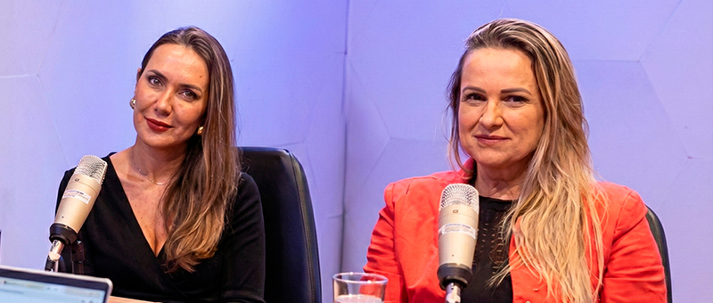 A juíza e a promotora  estão sentadas à mesa em um estúdio, com microfones posicionados à frente, como em uma gravação de podcast. Elas olham para a câmera com expressão serena e atenta; uma veste roupa preta e a outra, um blazer vermelho. Sobre a mesa há um copo com água e um computador