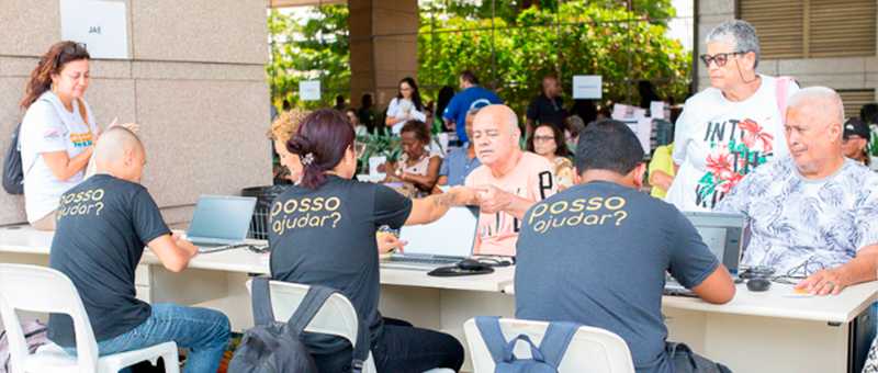 Imagem mostra idosos sentados em frente a uma mesa e sendo atendidos por pessoas, também sentadas, em frente a eles
