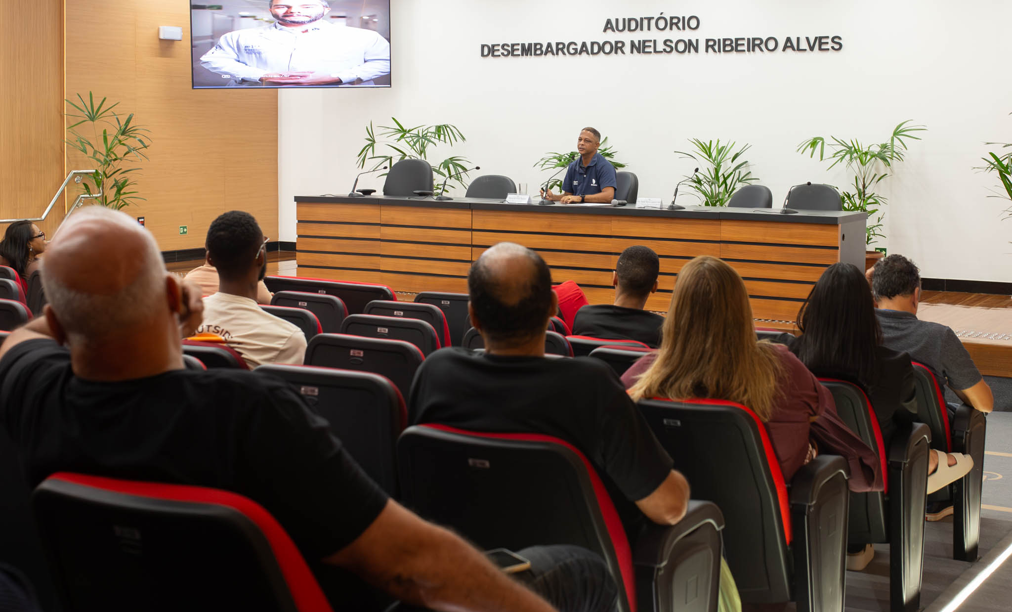 A imagem mostra um auditório durante uma palestra, com o público sentado de frente para o palco. Em primeiro plano, aparecem os participantes vistos de costas, distribuídos pelas poltronas estofadas nas cores preta e vermelha.  No palco, um homem de camisa polo azul está sentado à mesa principal, falando ao microfone. Ao fundo, há uma grande bancada com cadeiras vazias, microfones e vasos com plantas ornamentais. Na parede, destaca-se a inscrição "Auditório Desembargador Nelson Ribeiro Alves". No canto superior esquerdo, um telão exibe a imagem de um homem de jaleco branco, aparentemente participando da apresentação por vídeo. O ambiente é amplo, bem iluminado e possui acabamento em madeira nas paredes laterais.