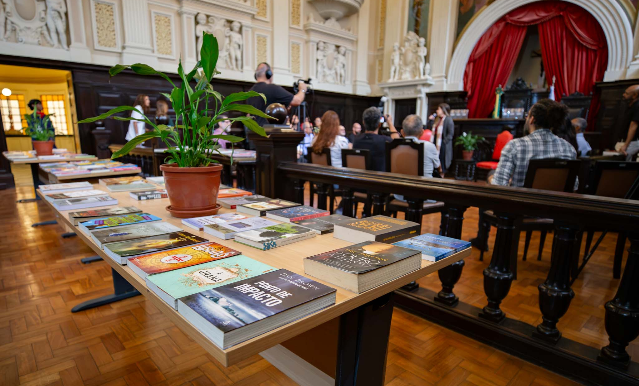 Em primeiro plano, uma mesa exibe dezenas de livros organizados lado a lado, com destaque para um vaso de planta ao centro, compondo um espaço destinado à troca de livros. Ao fundo, desfocado, um grupo de pessoas participa de uma atividade no salão histórico do Museu da Justiça. Um cinegrafista registra o evento enquanto uma mulher fala ao público. O ambiente é marcado por elementos arquitetônicos clássicos, como painéis ornamentados, piso de madeira, mobiliário escuro e um palco ao fundo com cortinas vermelhas, reforçando o caráter histórico e cultural do espaço.