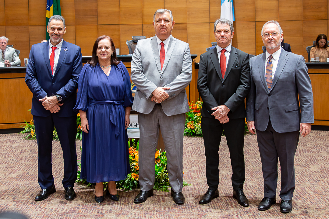 A imagem mostra cinco pessoas posando para uma foto oficial no Tribunal Pleno do TJRJ. Elas estão lado a lado, em pé, vestidas de forma formal. Da esquerda para a direita: um homem usando terno azul-marinho com gravata vermelha, sorrindo levemente; uma mulher com vestido azul escuro de mangas longas e colar, também sorrindo; um homem ao centro com terno cinza e gravata vermelha, com expressão mais séria; outro homem com terno preto e gravata vermelha, expressão neutra; e um homem de terno cinza claro com gravata estampada, usando óculos e com barba grisalha. Ao fundo, há uma mesa elevada com outras pessoas sentadas, bandeiras (incluindo a do Brasil) e um painel de madeira. Na parte frontal do espaço, há arranjos de flores em tons de laranja e verde.