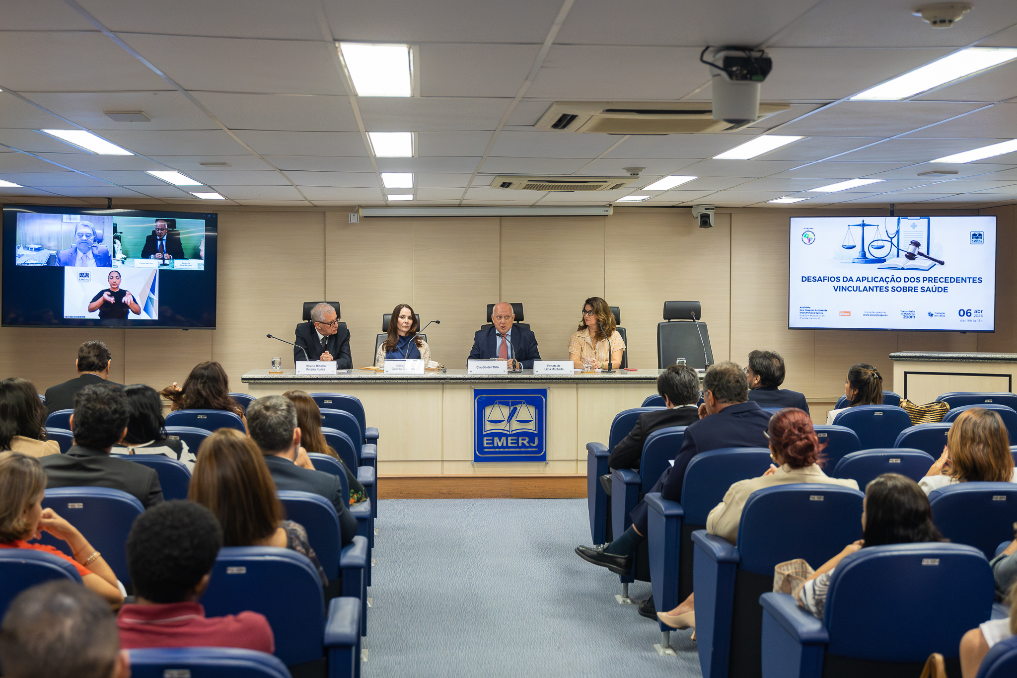A imagem mostra um auditório durante a realização de um evento institucional. Em primeiro plano, há o público sentado em poltronas azuis, voltado para a mesa principal. À frente, quatro palestrantes estão sentados atrás de uma bancada, cada um com microfone e identificação, participando de um painel de debate. Ao centro, um homem fala enquanto os demais escutam atentamente. O ambiente é bem iluminado e organizado, com aparência formal. Na parede ao fundo, há duas telas. À direita, um slide exibe o tema do evento: “Desafios da aplicação dos precedentes vinculantes sobre saúde”, acompanhado de elementos gráficos relacionados ao Direito, como uma balança da justiça. À esquerda, outra tela mostra participantes remotos e uma intérprete de Libras, indicando que o evento conta com transmissão ou participação híbrida. A mesa principal traz o símbolo da EMERJ.