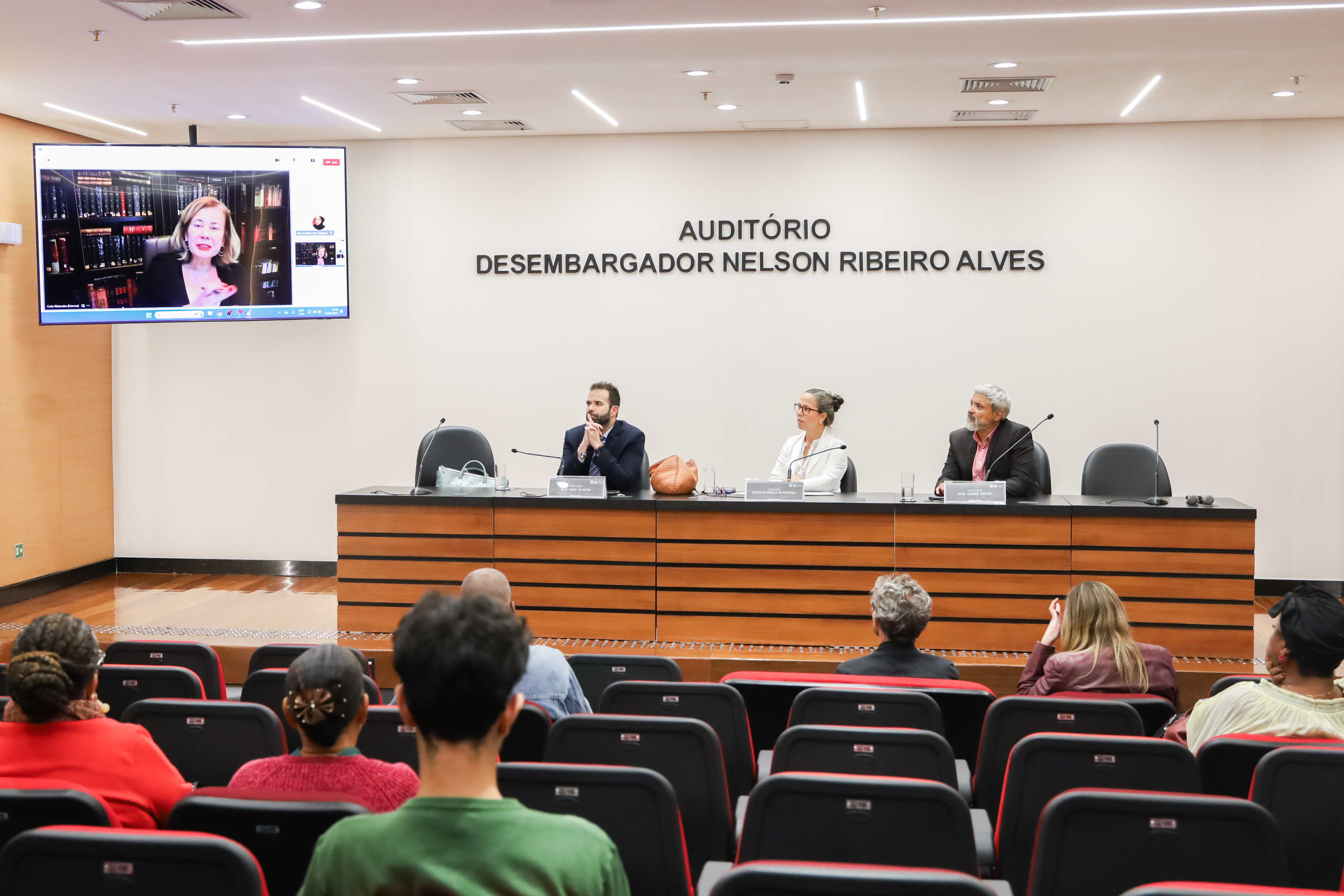 A imagem mostra o auditório cheio com três pessoas (dois homens e uma mulher) na mesa de debates e uma moça no telão.