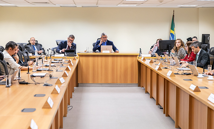 A imagem mostra uma reunião formal. A disposição é em formato de “U”, com mesas de madeira clara alinhadas dos dois lados e uma mesa central ao fundo, onde se encontra o desembargador Marcelo Anátocles que conduz a reunião. Todas as pessoas estão sentadas, a maioria com laptops ou documentos à frente.  À direita da imagem, uma bandeira do Brasil está posicionada no canto, 