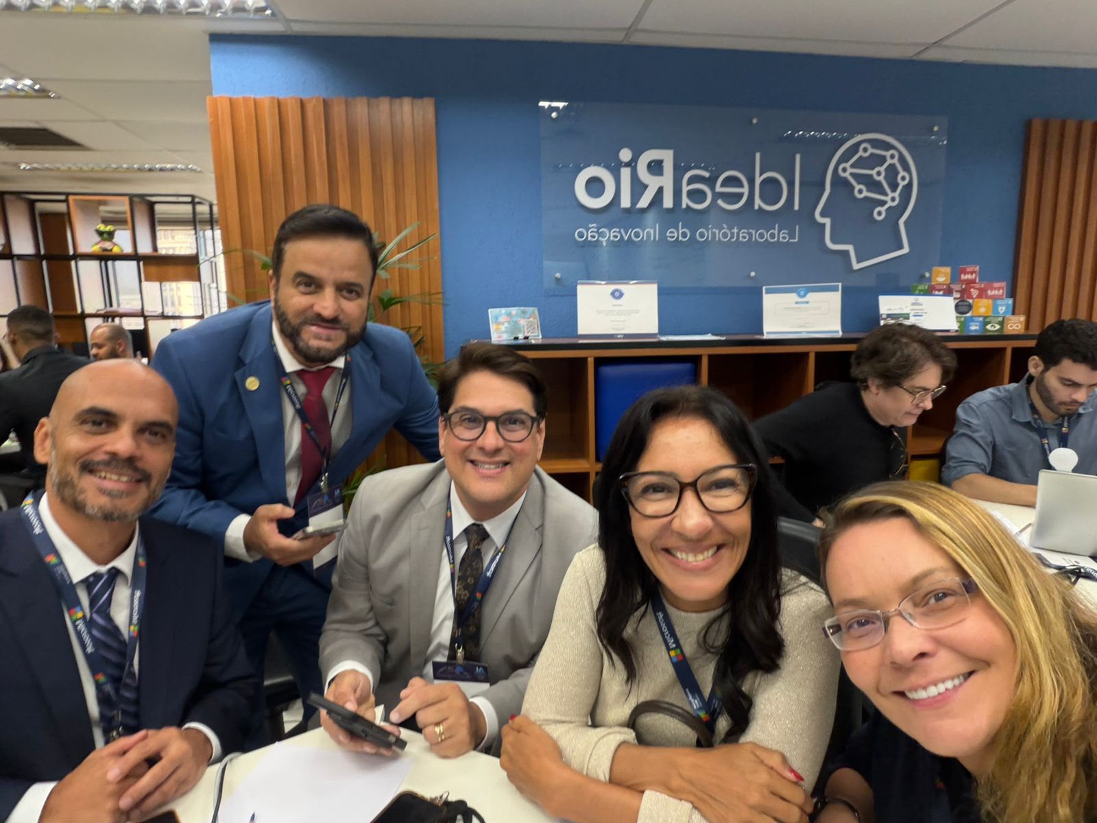 A foto apresenta cinco pessoas ao redor de uma mesa, três homens à esquerda e duas mulheres à direita.