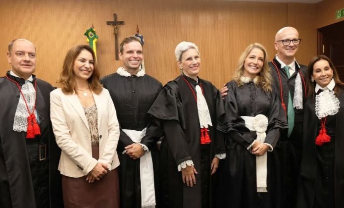 A imagem mostra um grupo de sete magistrados posando para uma foto em um ambiente interno e institucional. A maioria delas está vestindo togas pretas. Algumas togas têm detalhes em renda branca nas mangas e no peito, além de cordões vermelhos pendurados. No fundo, a parede é de madeira clara, e há uma cruz e símbolos do Brasil.