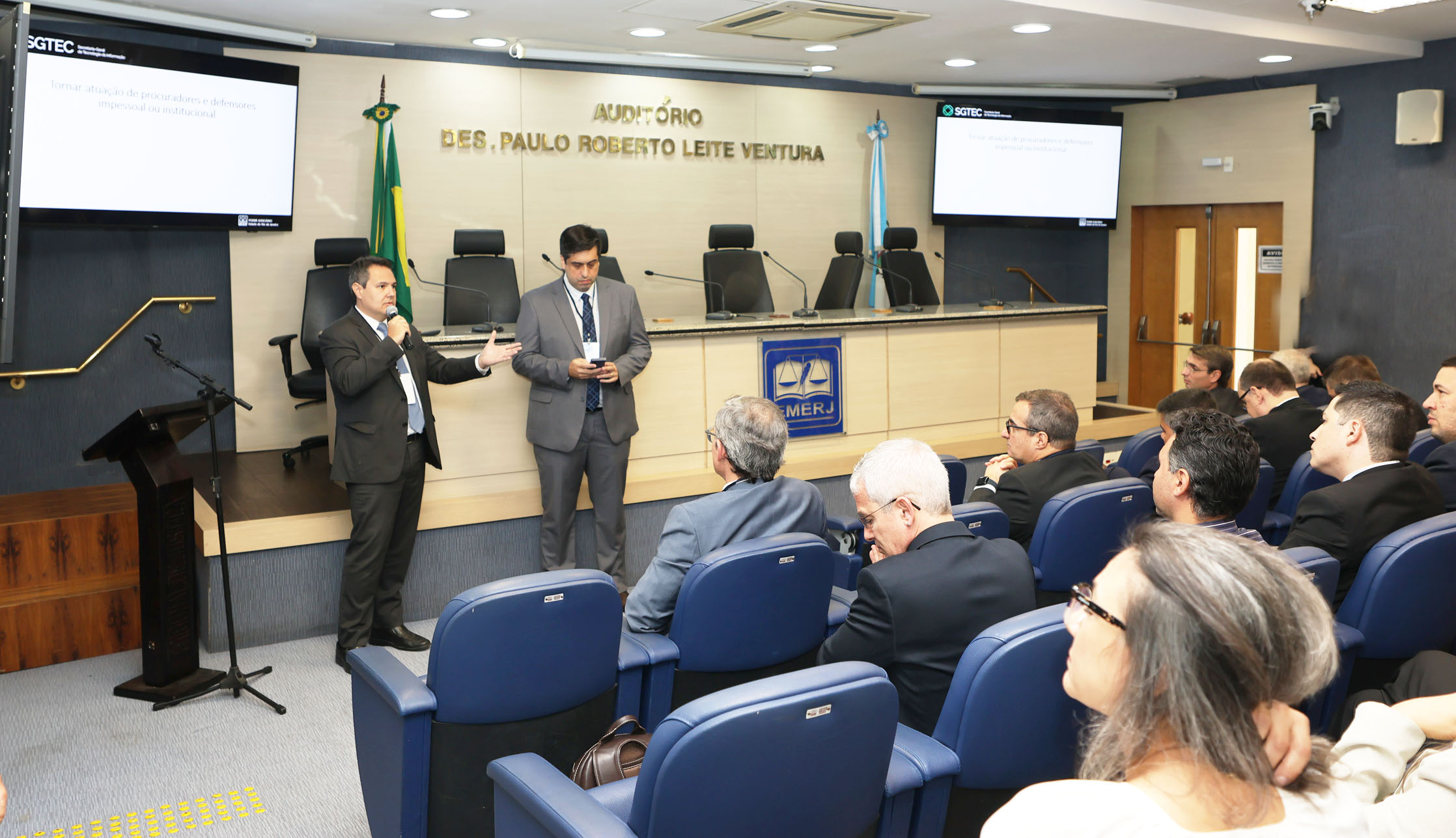 A imagem mostra o Auditório Desembargador Paulo Roberto Leite Ventura com dois homens em pé na frente apresentando uma palestra