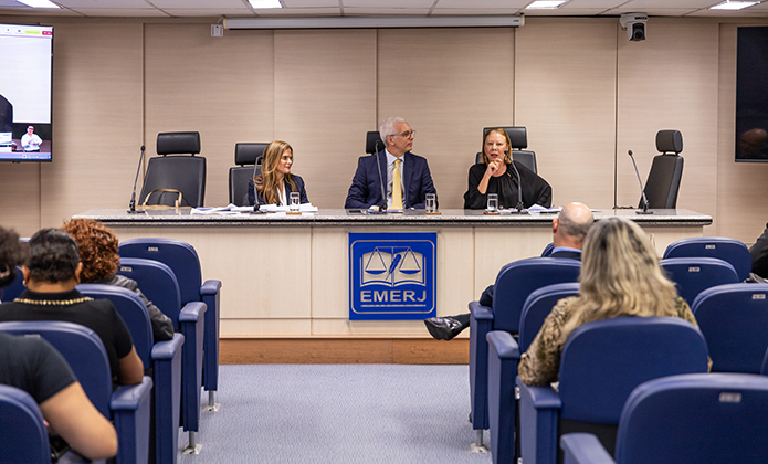 A imagem mostra o auditório da EMERJ (Escola da Magistratura do Estado do Rio de Janeiro), onde ocorre a palestra. Três pessoas estão sentadas à mesa principal, em posição de destaque, com microfones e papéis à frente. O desembargador Wagner Cinelli (no centro) veste terno e gravata, enquanto as duas mulheres (juíza Luciana Fiala e desembargadora Maria Angélica Guerra) usam roupas formais. Uma delas, a desembargadora, está falando ao microfone.  A plateia, composta por várias pessoas, está sentada em cadeiras azuis voltadas para a mesa principal. O ambiente é bem iluminado, com paredes claras e um telão ao fundo transmitindo a imagem do evento.