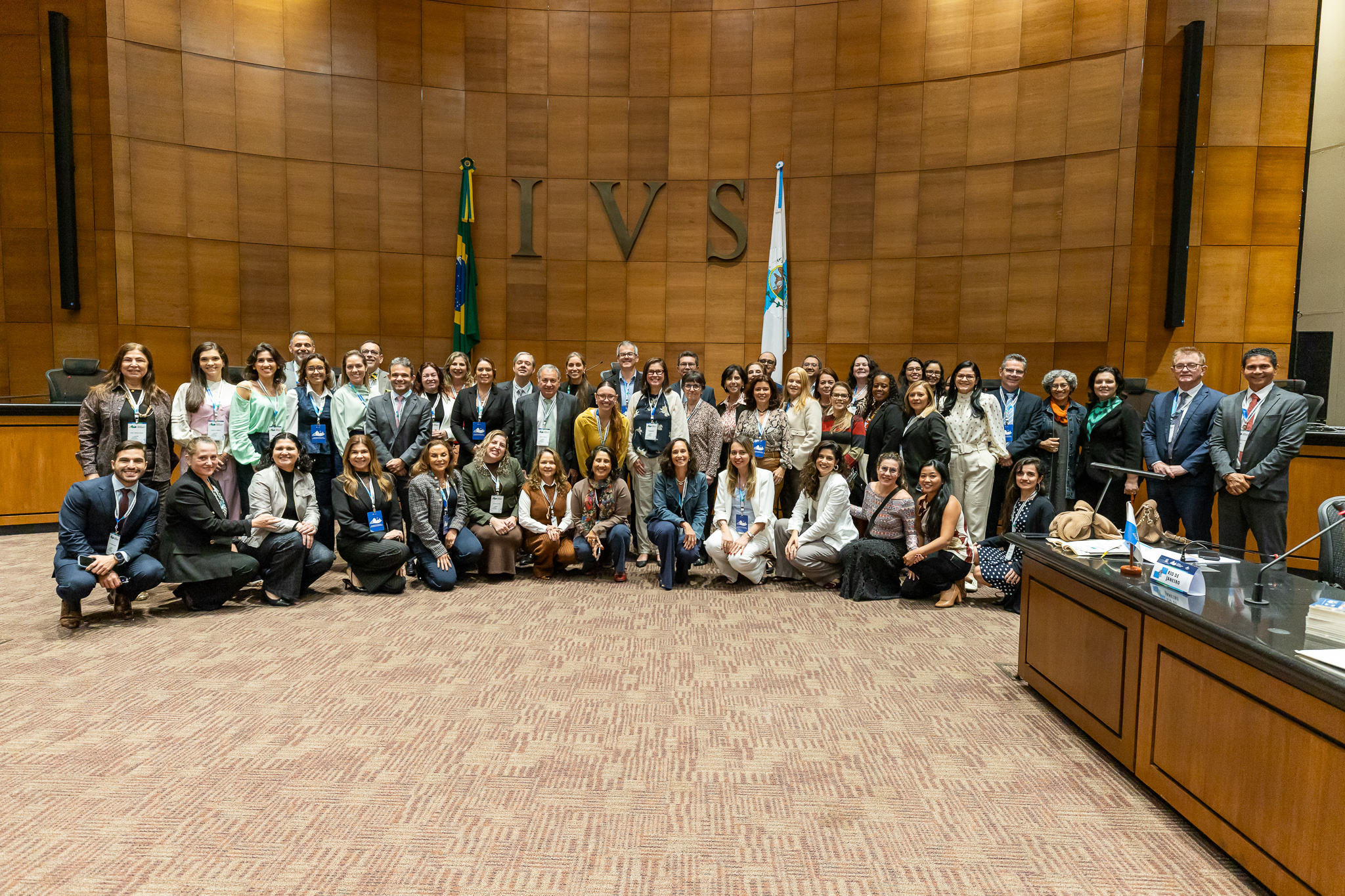 A imagem mostra uma fotografia de grupo realizada em um auditório institucional. Cerca de cinquenta pessoas posam para a foto, distribuídas entre fileiras em pé e agachadas na parte frontal. A maioria veste trajes sociais ou esporte fino, e muitos utilizam crachás de identificação pendurados no pescoço. Ao fundo, destaca-se uma parede revestida de madeira com a inscrição em letras grandes “IVS”, termo em latim associado ao conceito de Direito e Justiça. Também são visíveis as bandeiras do Brasil e do Estado do Rio de Janeiro, reforçando o caráter oficial do ambiente. À direita da imagem aparece uma mesa de autoridades, equipada com microfones e identificação institucional. 