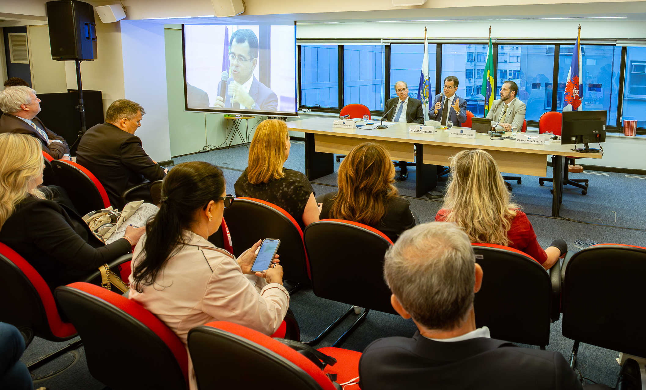 A imagem mostra um auditório de pequeno porte durante um evento institucional. Em primeiro plano, há pessoas sentadas em cadeiras acolchoadas pretas com detalhes em vermelho, de costas para a câmera, acompanhando atentamente a apresentação. Uma mulher, à esquerda, utiliza o celular, enquanto outras pessoas mantêm o olhar voltado para a frente. Na parte frontal da sala, três homens estão sentados atrás de uma mesa, participando de um painel de debate. Um deles fala ao microfone e gesticula com as mãos, indicando que está conduzindo ou respondendo a uma questão. Sobre a mesa, há copos de água, microfones e placas de identificação. Atrás dos palestrantes, há bandeiras — incluindo a do Brasil — e uma grande janela que permite a entrada de luz natural. À esquerda, um telão exibe a imagem ampliada de um dos participantes falando ao microfone, reforçando o caráter formal e organizado do evento.