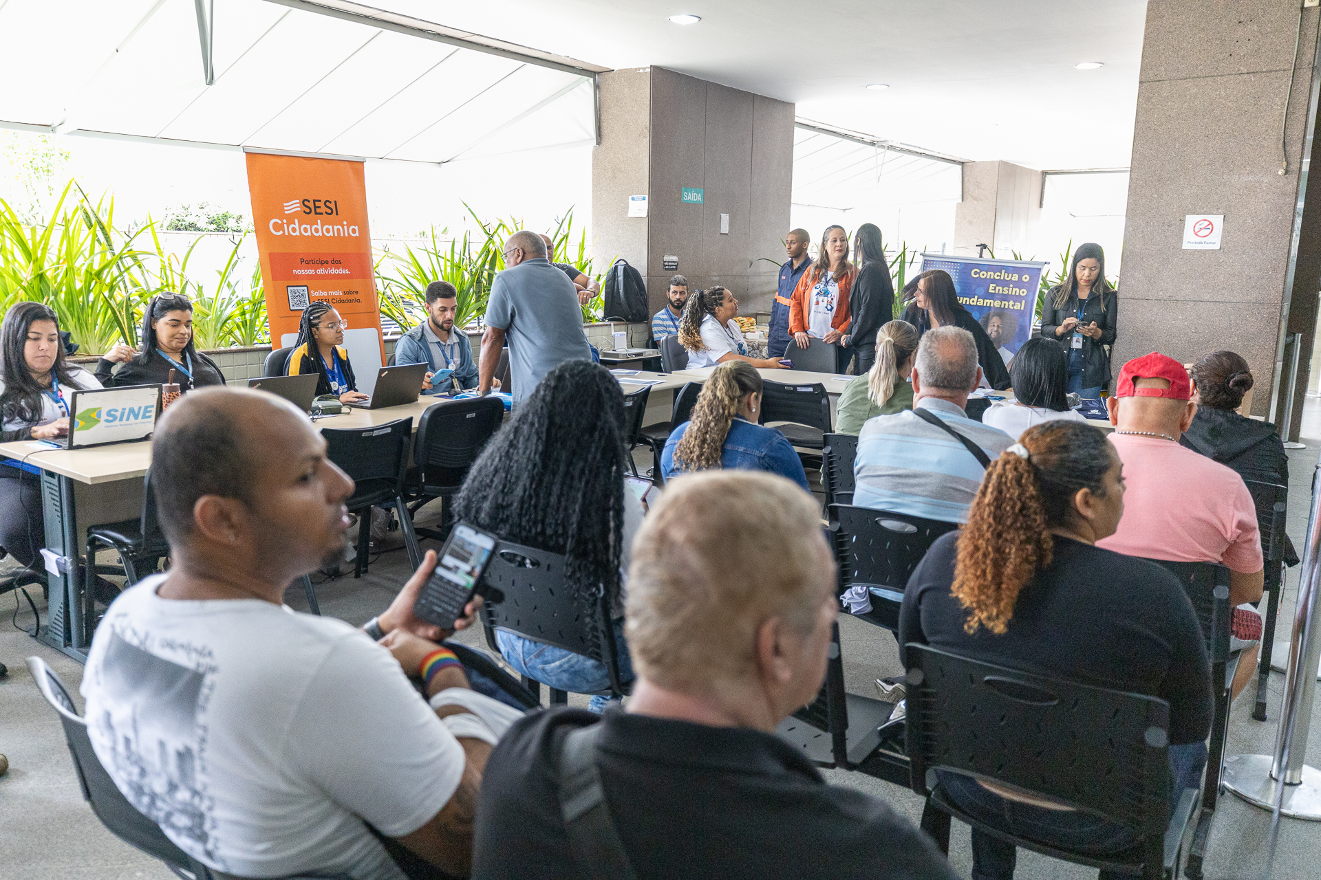 Imagem do Fórum de Madureira durante ação de empregabilidade. Pessoas de costas sentadas em cadeiras aguardando atendimento. Ao fundo, funcionários do fórum sentados em mesas. 