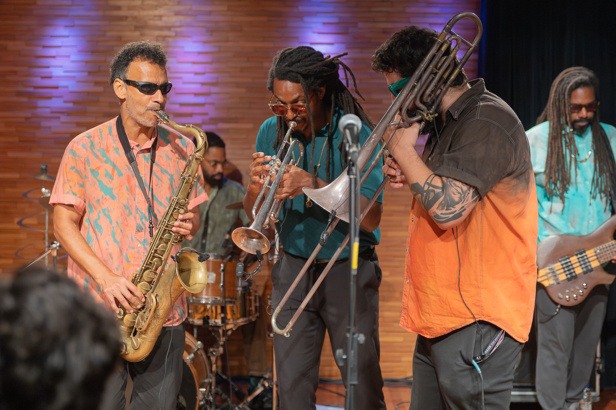 A imagem mostra uma apresentação musical ao vivo em um ambiente fechado, com clima intimista e iluminação quente. No centro da cena, três músicos tocam instrumentos de sopro próximos a um microfone: À esquerda, um homem usa óculos escuros e camisa estampada em tons alaranjados enquanto toca um saxofone, com postura concentrada. No meio, outro músico, com cabelos em dreadlocks e óculos, toca um trompete, inclinado levemente para frente, em sintonia com os demais. À direita, um terceiro músico, com barba e tatuagem no braço, veste uma camisa em degradê alaranjado e toca um trombone, com o instrumento estendido em movimento. Ao fundo, outros integrantes da banda acompanham a apresentação: um baterista aparece parcialmente atrás dos músicos principais, tocando a bateria. À direita, um baixista com dreadlocks toca um baixo elétrico, completando a formação. O cenário possui uma parede de madeira com iluminação suave em tons de roxo e azul, criando uma atmosfera acolhedora e artística.