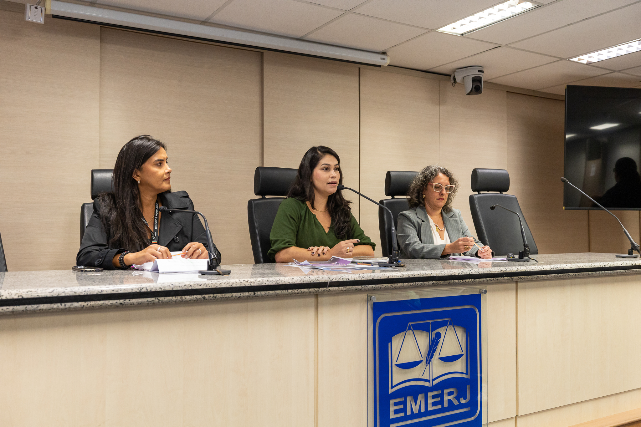 Foto mostra três mulheres, sentadas em mesa de auditório, sendo uma delas falando ao microfone, indicando que está proferindo uma palestra