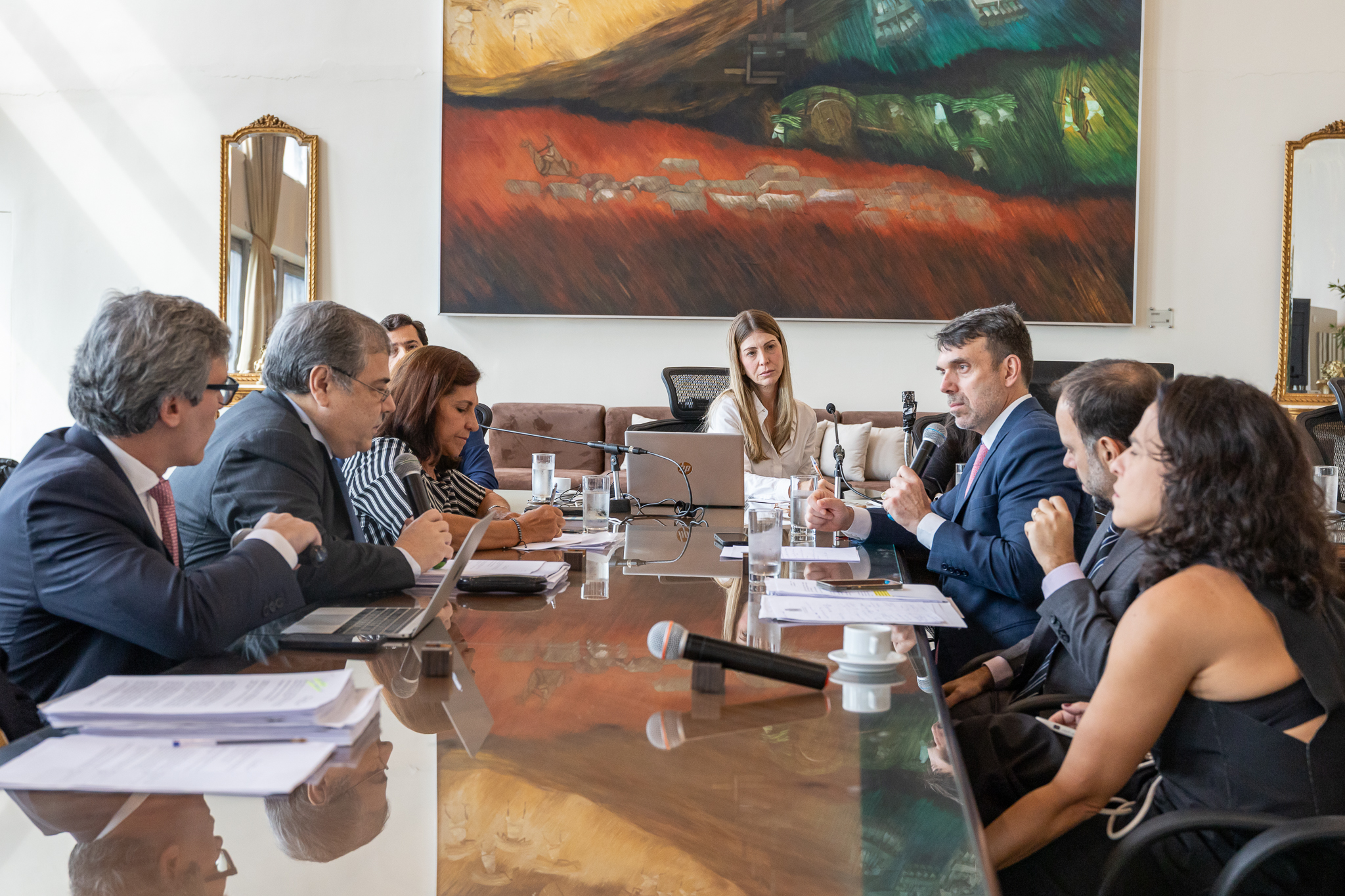 Foto de audiência pública realizada em salão grande, com homens e mulheres sentados em volta de uma mesa, que tem em sua cabeceira uma mulher, a juíza que preside a audiência. Ao fundo da sala se destaca um grande quadro de pintura