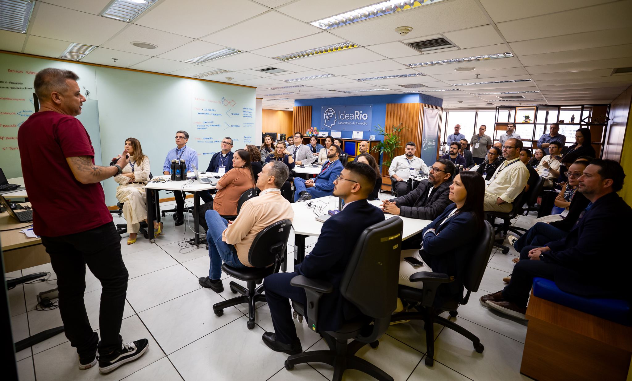 A imagem mostra uma apresentação realizada em um ambiente corporativo voltado à inovação. Em primeiro plano, um homem de camiseta vermelha está em pé, falando para um grupo de participantes sentados. Ele gesticula enquanto apresenta o conteúdo, diante de uma tela e de um quadro branco com anotações. O público é composto por cerca de 30 pessoas, entre homens e mulheres, distribuídas em cadeiras ao redor de mesas de trabalho. Os participantes observam atentamente o palestrante, alguns com notebooks, celulares e materiais de apoio sobre as mesas. Ao fundo, destaca-se um espaço identificado pela marca IdeaRio – Laboratório de Inovação, com painéis azuis, divisórias de madeira e elementos decorativos que reforçam o ambiente colaborativo.