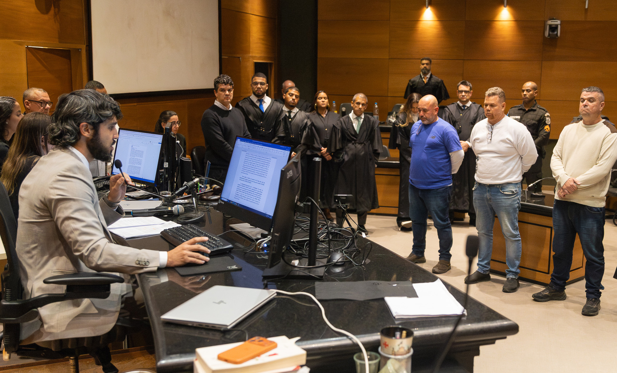 A foto mostra a imagem do plenário durante a leitura da sentença com o juiz à esquerda e os três réus à direita.