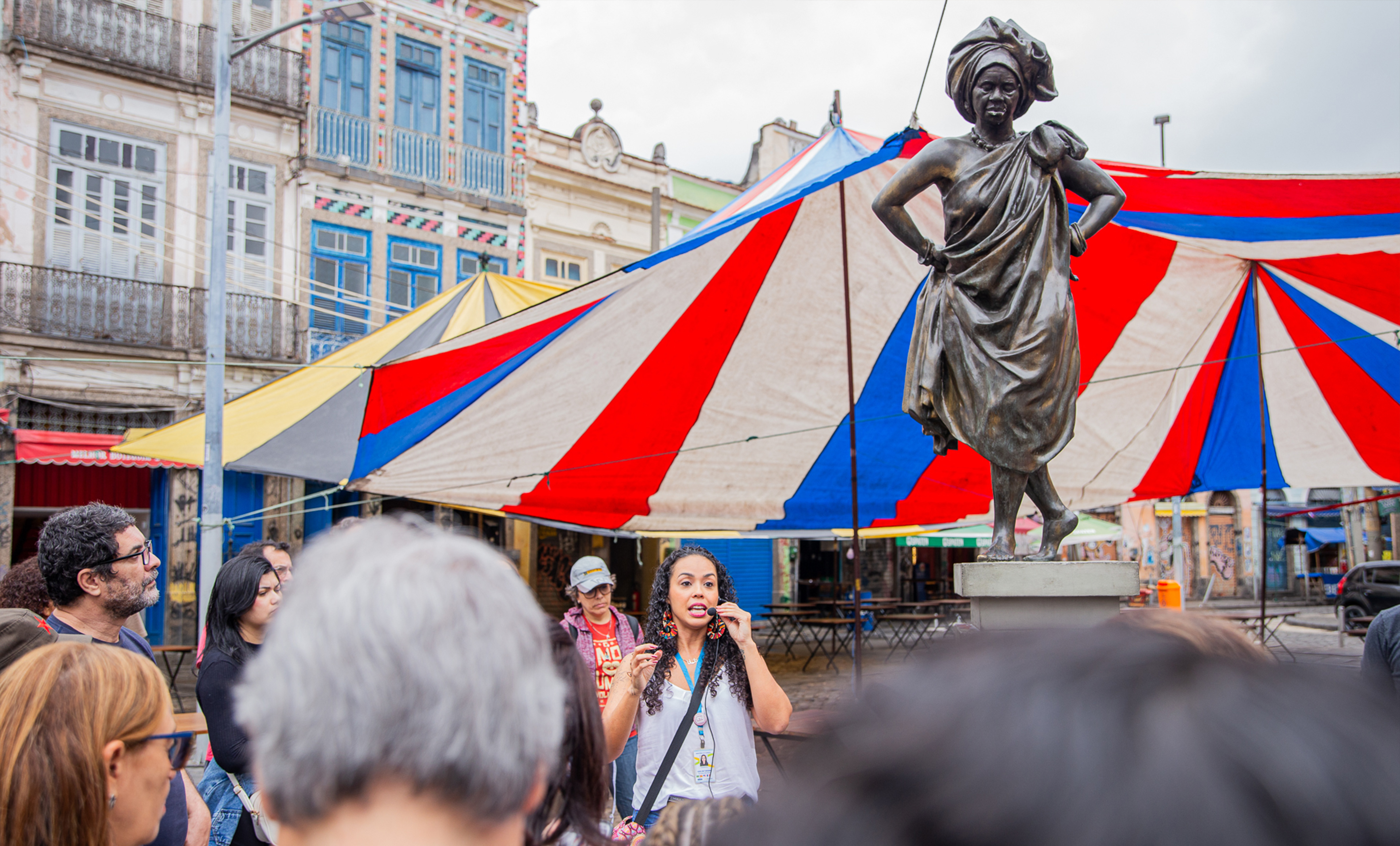 Imagem mostra um grupo de pessoas reunidas em uma praça durante uma atividade guiada ao ar livre. Em primeiro plano, aparece parte do público observando uma mulher que fala ao microfone, aparentemente conduzindo a visita ou apresentação. Ao lado dela, destaca-se uma estátua de mulher negra em bronze, com uma das mãos na cintura e vestimentas tradicionais, posicionada sobre um pedestal.  Ao fundo, há grandes tendas listradas nas cores vermelha, azul e branca, além de prédios antigos com fachadas históricas e detalhes arquitetônicos coloridos, compondo um cenário urbano e cultural. 