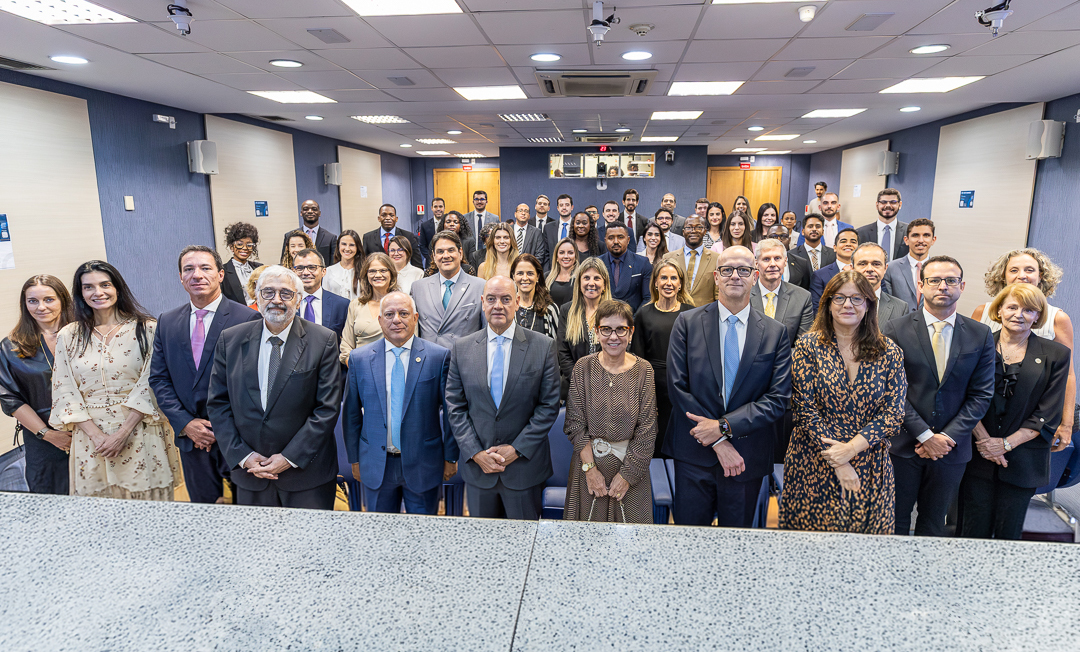 A imagem mostra uma foto oficial de grupo em um auditório institucional, reunindo um grande número de pessoas dispostas em várias fileiras. A maioria veste trajes formais, como ternos, gravatas, vestidos e roupas sociais, o que indica um evento solene, possivelmente uma cerimônia, curso ou ato oficial.