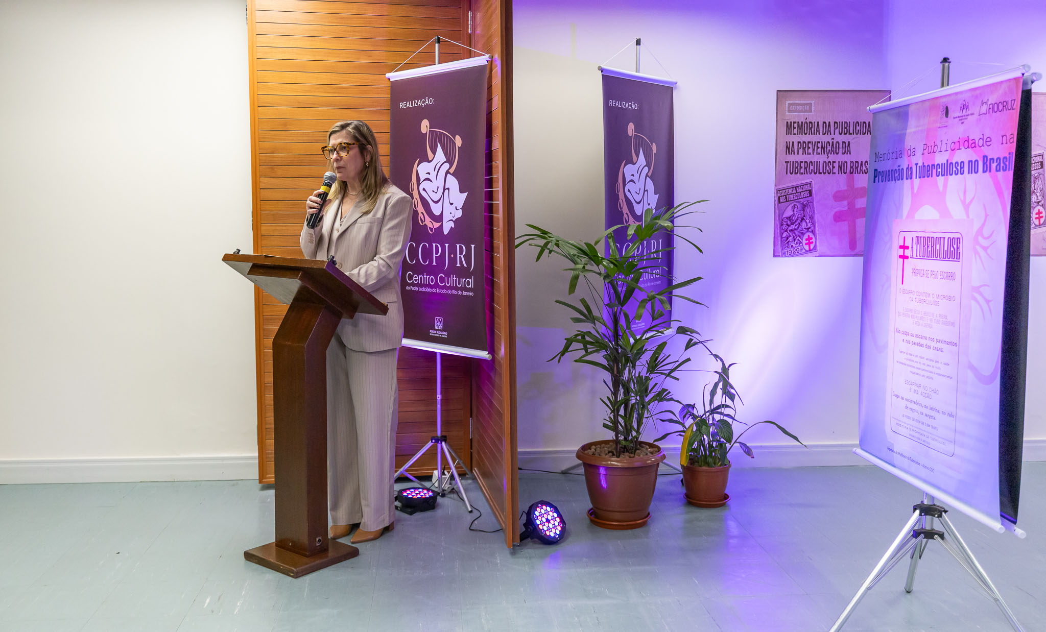 A imagem mostra a abertura de uma exposição em um espaço interno. Em primeiro plano, a diretora do CCPJ, Ana Paula Teixeira Delgado, está em pé atrás de um púlpito de madeira, falando ao microfone e olhando para o público. Ela veste roupas claras. Ao fundo, há um banner institucional e painéis expositivos sobre a história da prevenção da tuberculose no Brasil. À direita da imagem, destaca-se um painel com a reprodução de um cartaz histórico, montado sobre um tripé. Vasos com plantas ornamentais complementam o ambiente, que conta com iluminação em tons de roxo.