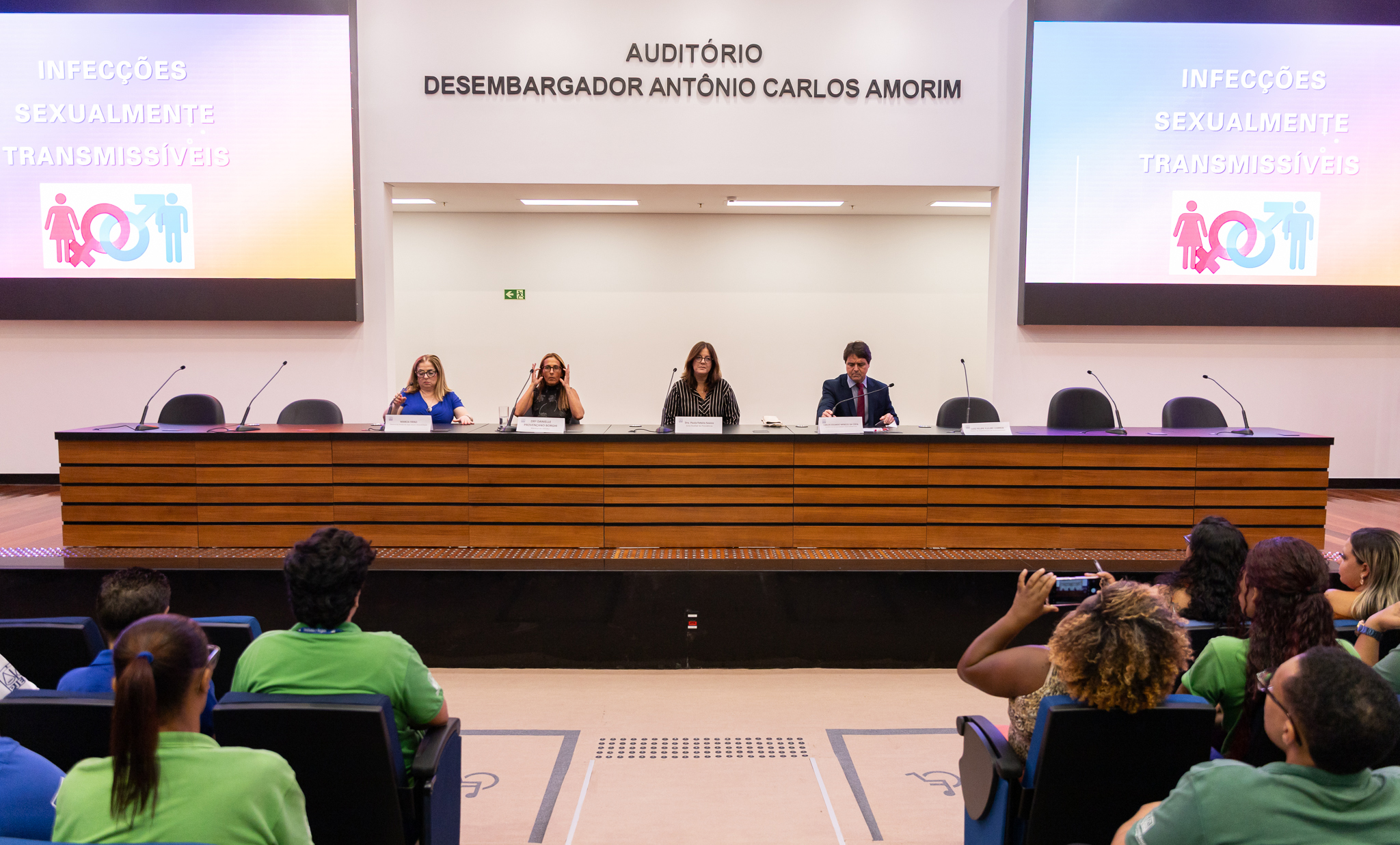 Palestrantes sentados à mesa em auditório diante de plateia de jovens que são informados sobre IST