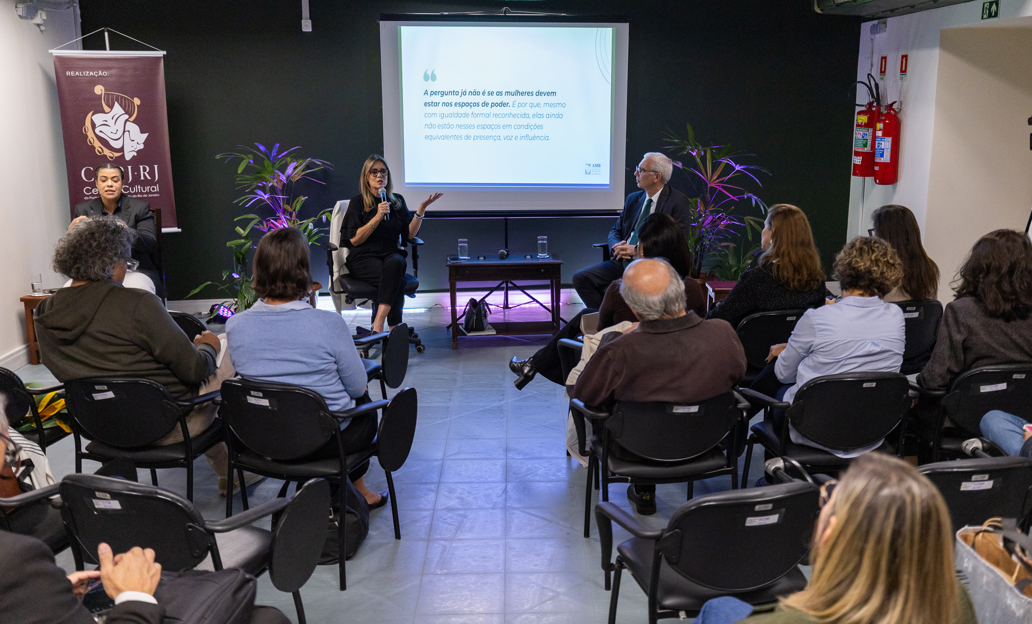 A imagem registra uma palestra ou mesa de debate realizada no Centro Cultural do Poder Judiciário do Estado do Rio de Janeiro (CCPJ-RJ). O evento acontece em uma sala de auditório, com o público acomodado em cadeiras voltadas para os palestrantes. Ao centro, uma mulher sentada segura um microfone e fala ao público, enquanto faz um gesto com uma das mãos. Ao seu lado, um homem de terno acompanha a apresentação. À esquerda, outra participante também está sentada, compondo a mesa de debate. Ao fundo, uma tela de projeção exibe uma citação sobre a presença e a influência das mulheres em espaços de poder. O ambiente é decorado com plantas ornamentais iluminadas por luzes coloridas, criando um cenário acolhedor para a discussão. Um banner do CCPJ-RJ está posicionado à esquerda da tela. Na plateia, homens e mulheres de diferentes idades acompanham atentamente a apresentação. 