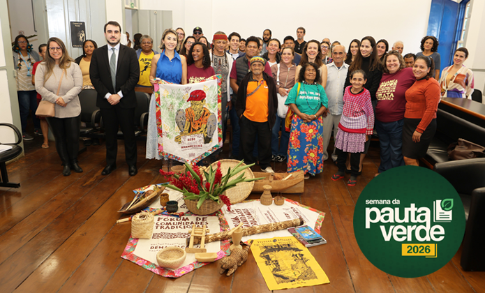 A imagem mostra um grupo numeroso de pessoas reunidas em uma sala para uma fotografia coletiva durante um evento da Semana da Pauta Verde 2026. Os participantes, de diferentes idades, estão posicionados em pé, lado a lado, sorrindo para a câmera. No centro da composição, algumas pessoas seguram um estandarte colorido, enquanto, no chão, estão expostos diversos objetos ligados à cultura tradicional e aos povos originários e comunidades tradicionais, como cestos de palha, instrumentos artesanais, sementes, livros, publicações e arranjos com flores e folhagens. Também é possível observar faixas e materiais informativos dispostos sobre tecidos estampados. O ambiente possui piso de madeira, paredes claras e mobiliário de auditório ao redor. À direita da imagem, destaca-se a identidade visual do evento, com o logotipo da Semana da Pauta Verde 2026.