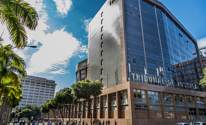 A imagem mostra a fachada lateral do Tribunal de Justiça do Rio de Janeiro. O prédio é alto, com arquitetura moderna, revestido em vidro espelhado e estrutura em tons de bege e marrom. A parte superior reflete o céu azul com nuvens brancas, criando contraste entre o concreto urbano e o ambiente natural. Na base do edifício, há uma área com janelas retangulares alinhadas e uma marquise onde se lê a inscrição “TRIBUNAL DE JUSTIÇA” em letras metálicas. À frente, uma calçada ampla com circulação de pedestres indica movimento cotidiano na região. À esquerda da imagem, aparecem palmeiras e árvores que emolduram a cena, além de outro prédio ao fundo.