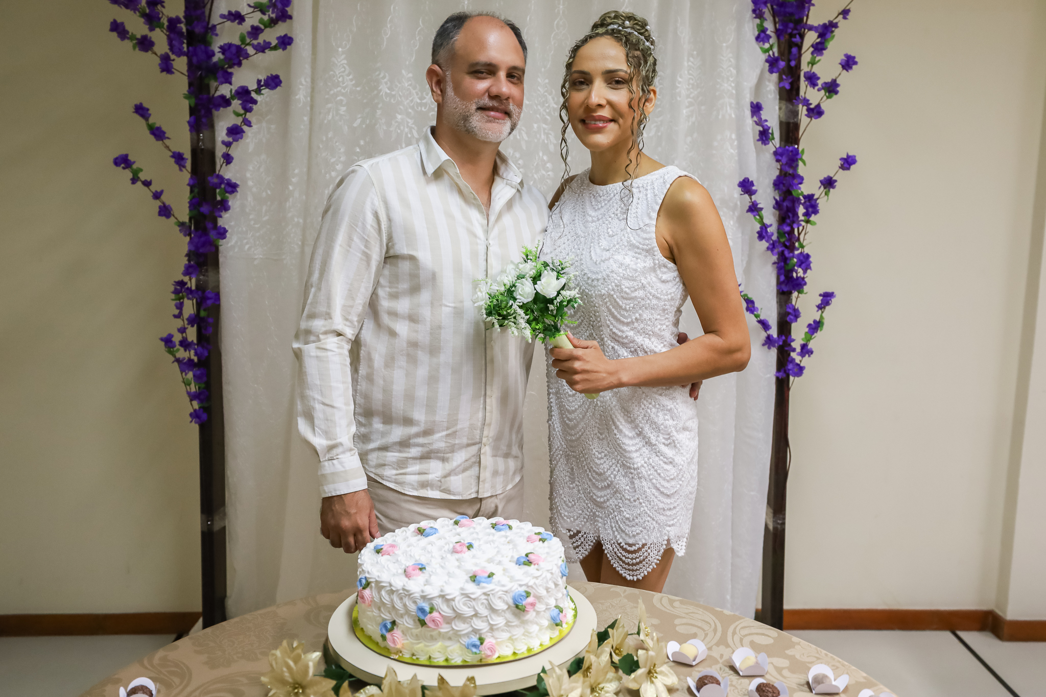 Foto de casal de noivos, vestidos de branco, em frente à mesa com bolo de casamento e docinhos
