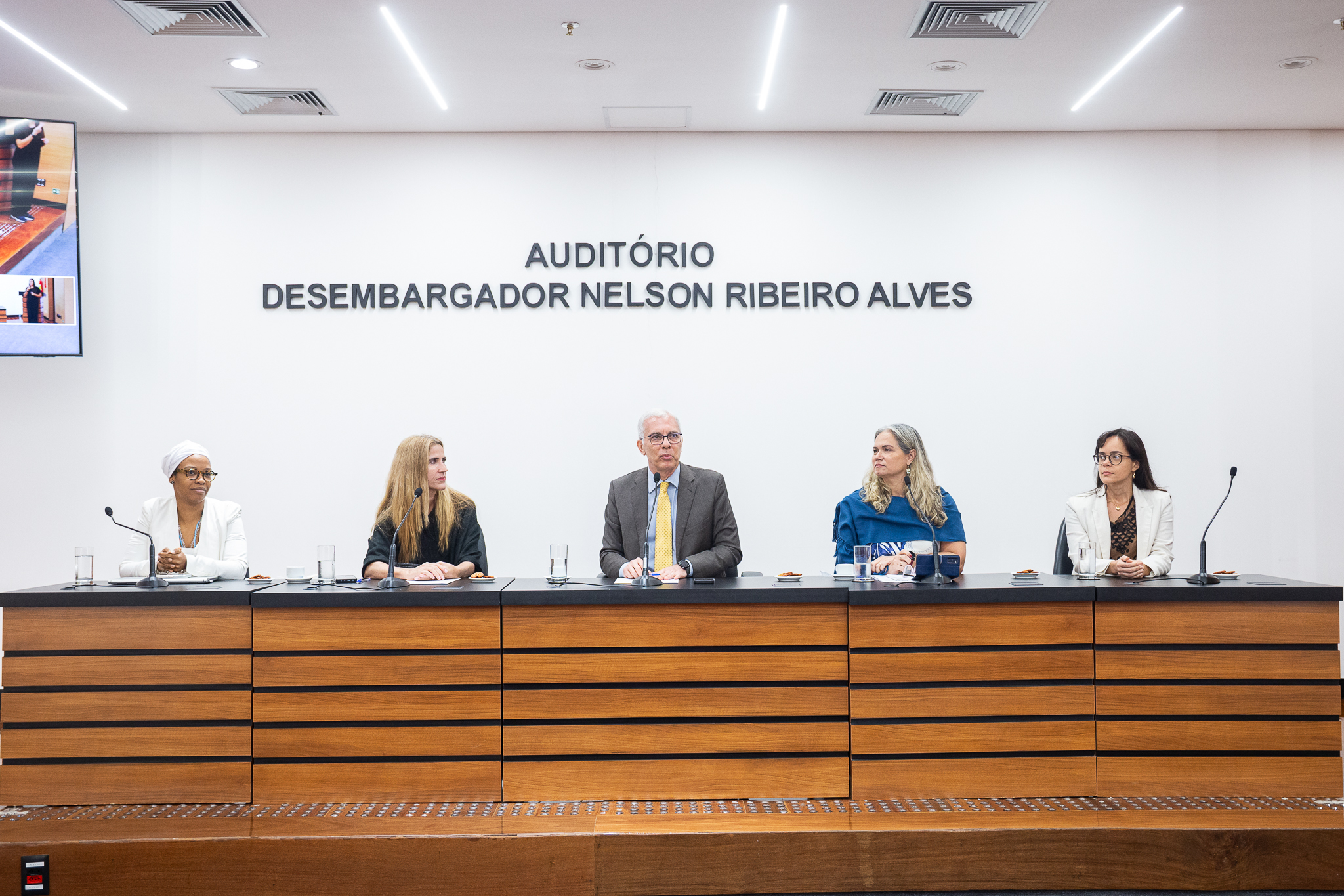 A imagem mostra uma mesa de debate realizada em um auditório, com cinco participantes sentados lado a lado atrás de uma bancada equipada com microfones. Ao centro, o desembargador Wagner Cinelli, um homem de terno escuro e gravata amarela e que fala ao microfone, com postura ereta e expressão séria. Os demais participantes — quatro mulheres — estão atentos, voltados para ele ou para o público, demonstrando escuta e participação no debate. À esquerda, uma das mulheres usa roupas claras e um turbante branco, enquanto as outras vestem trajes formais em tons neutros. Sobre a mesa, há copos com água, xícaras de café e pequenos pratos. Ao fundo, na parede branca, está escrito “Auditório Desembargador Nelson Ribeiro Alves”.
