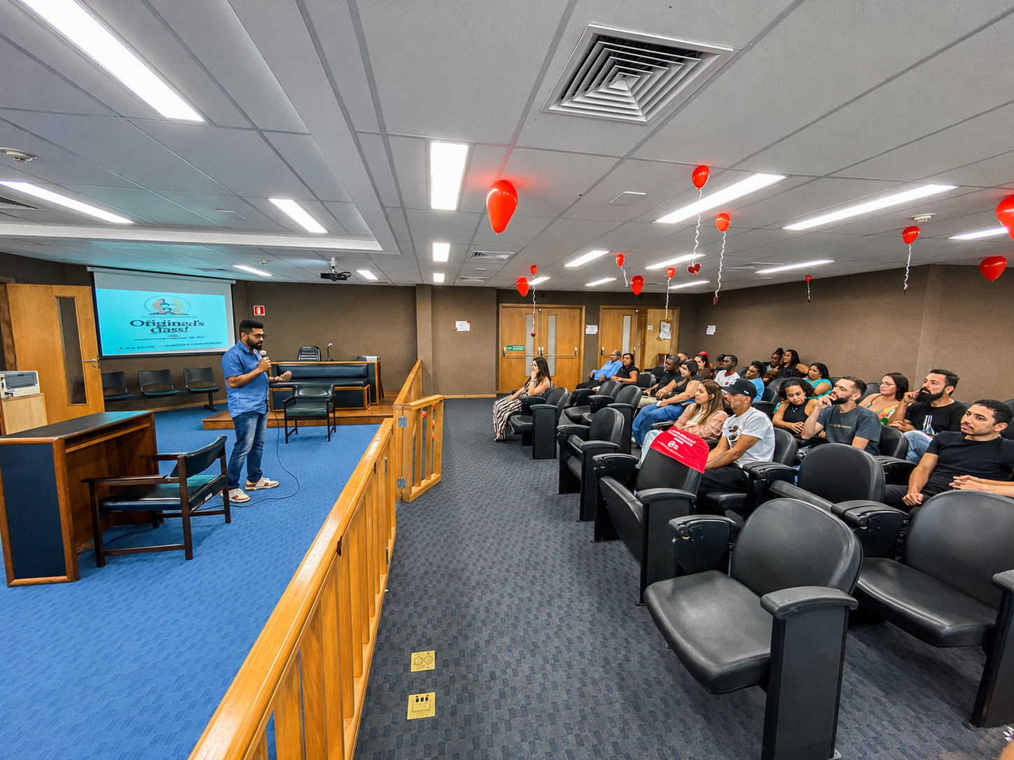 A imagem mostra um auditório durante uma palestra ou atividade educativa. À esquerda, um homem veste camisa polo azul e calça jeans, segura um microfone e fala ao público próximo ao palco. Atrás dele, um telão exibe uma apresentação com o título "Oficinas Grátis!", acompanhado de uma ilustração de um casal e de informações sobre o curso. À direita, cerca de 20 participantes estão sentados em poltronas pretas, voltados para o palestrante. O grupo é composto por homens e mulheres de diferentes idades, que acompanham atentamente a apresentação. O ambiente possui piso acarpetado, palco elevado com mobiliário semelhante ao de uma sala de audiências, incluindo mesa, cadeiras e bancada. O teto é decorado com balões vermelhos em formato de coração, presos por fitas, reforçando o caráter temático da atividade.   