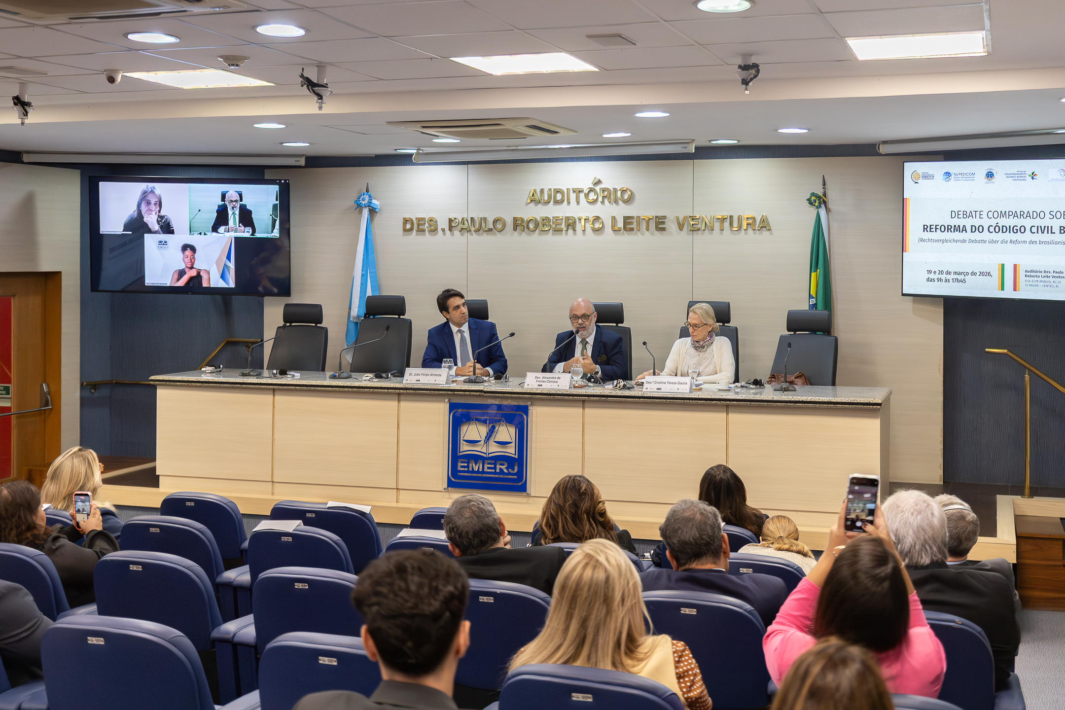 A imagem mostra um evento institucional realizado em um auditório, com uma mesa de autoridades à frente e público sentado na plateia. Ao fundo, na parede, está escrito em letras douradas: “AUDITÓRIO DES. PAULO ROBERTO LEITE VENTURA”. À frente, três participantes estão sentados na mesa principal, cada um com microfone, copo de água e identificação. No centro está o desembargador Alexandre de Freitas Câmara, um homem mais velho, de barba grisalha e óculos, veste terno e gravata e fala ao microfone, gesticulando levemente com as mãos, indicando que está conduzindo ou participando ativamente do debate. À esquerda dele, o advogado João Felipe Almeida, um homem mais jovem, de terno azul, observa com atenção, voltado para o orador. À direita, a desembargadora Cristina Tereza Gaulia, uma mulher de cabelos claros, usando óculos e roupas claras, lê ou consulta um documento sobre a mesa. Na parte frontal da mesa, aparece o símbolo da EMERJ (Escola da Magistratura do Estado do Rio de Janeiro). À esquerda da imagem, um telão exibe três participantes em videoconferência, sugerindo que o evento ocorre em formato híbrido. À direita, outro telão apresenta o tema do encontro, indicando tratar-se de um debate comparado sobre a reforma do Código Civil, com data e horário informados.